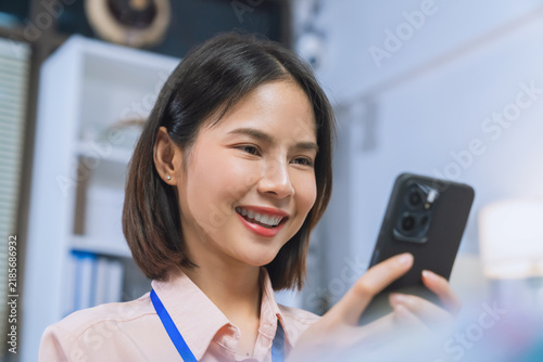 Smiling young Asian businesswoman using smartphone while working in office, mobile communication and business technology concept.