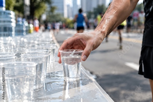 A runner grabs a cup of water from a table set up along a race course, with other runners in the background.