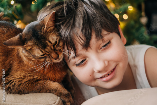 Kid boy teanager 12-years old with his pet red fluffy cat nearby decorated christmas tree closeup portrait somali breed cats
on the xmas hollidays at cozy home indoors lifestyle headshot