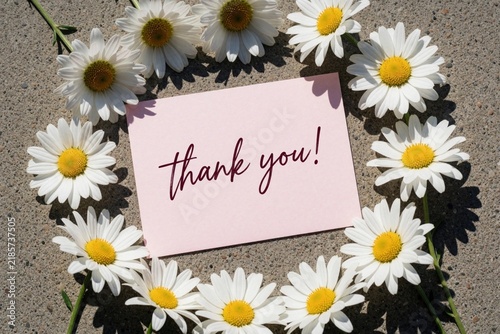 White daisy flowers form circular frame around pink card with thank text. Minimalist floral border on grey stone background. Beautiful chamomile blossom decoration for greeting card, wedding invite