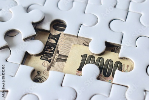 Fotografía High-angle close-up of Japanese yen banknotes visible through a missing piece in a white jigsaw puzzle, illustrating financial solution and hidden wealth concepts