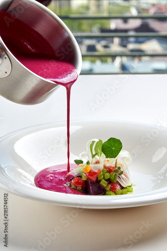 Pouring bright pink beetroot cream soup into a white bowl garnished with fresh greens and microgreens. High-end gourmet dining.