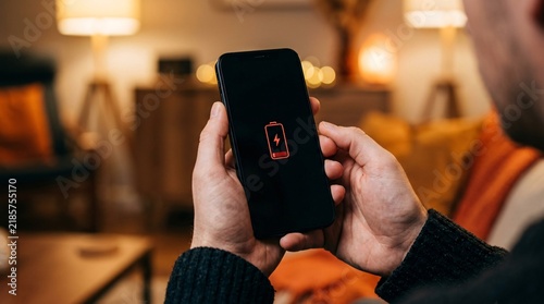 Man holding a smartphone displaying a red low battery warning icon on the screen in a living room.