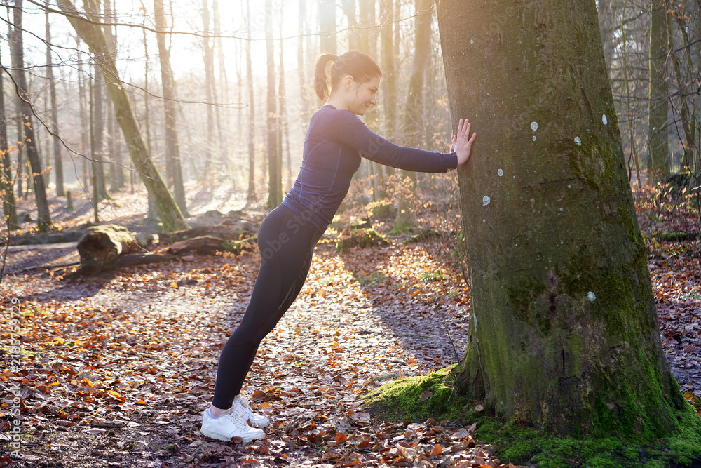 Obraz premium Athletic young sporty woman doing stretching exercises on a tree in forest on a sunny winter morning