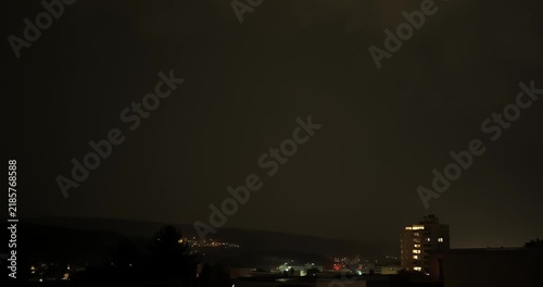 Frequent lightning flashes illuminate dark storm clouds above a distant apartment building and hillside village at night