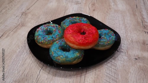 A set of four blue frosted donuts with colorful sprinkles is displayed on a black plate. The donuts are sitting on a wooden table. Someone's hand can be seen moving in the frame, suggesting an upcomin