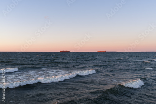 Aerial drone view of the choppy Baltic Sea featuring dynamic waves and two general cargo ships anchored on the distant horizon during a serene sunset.