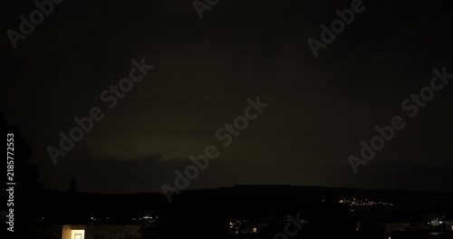 Heavy night storm clouds with frequent lightning flashes illuminating a small hillside village and residential area.