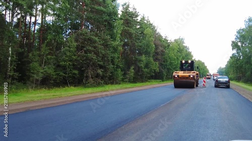 Construction work taking place on a highway surrounded by trees on both sides. Vehicles are moving through the area while workers and machines are busy with road repairs. This location is a rural area