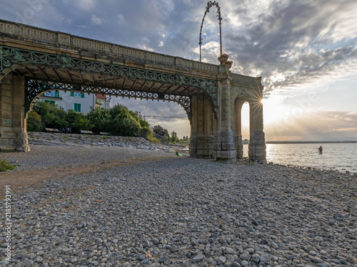 Obraz na plátně View from under the historic Schlosssteg pier at Lake Constance during sunrise, showcasing low water levels on a pebbled beach