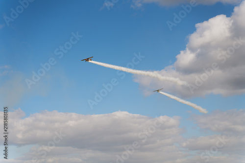 Two small stunt airplanes performing in an air show, leaving long white smoke trails against a blue sky with dramatic cumulus clouds.