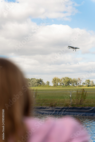 A view from behind a spectator watching a small single-engine airplane flying low over a green airfield and water reservoir under a cloudy blue sky.