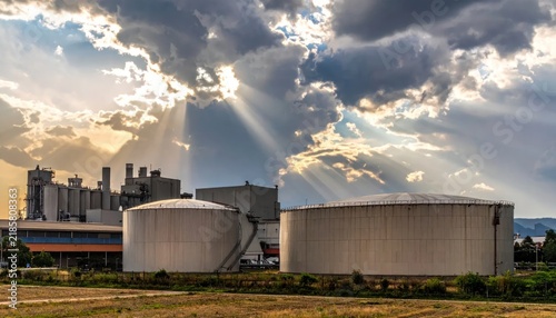 Industrial complex with large storage tanks under dramatic cloudy sky with sunbeams.