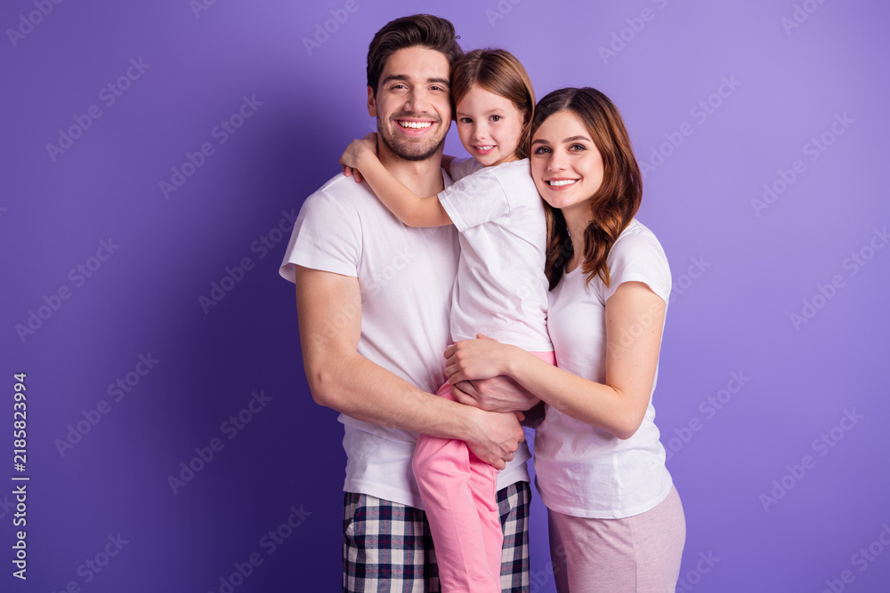 Fototapeta premium Smiling young family of mother, father and daughter hugging in casual pajamas against a purple studio background showing love and togetherness