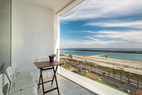 Modern balcony with white chairs and wooden table overlooking beach, ocean, blue sky, clouds, glass railing, potted plant, relaxing coastal resort setting, with balcony, chair, table, wooden detail