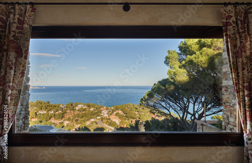 Scenic ocean view through interior window with foreground tree, coastal hills, calm horizon, and clear blue sky creating peaceful seaside composition, with ocean, window, view, coast detail