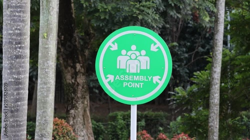 Green circular sign marks assembly point. White arrows point inward, guiding people. Three human figures shown, suggesting group meeting. Background features trees and orange flowering bushes