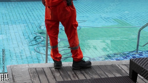 Worker in orange suit cleans swimming pool. He uses long pole to scrub pool surface. Pool water appears green and murky. Metal ladder sits beside tiled edge. Worker wears bright cap and sturdy boots