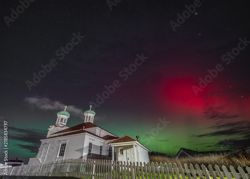 View of the historic Holy Ascension of Our Lord Cathedral illuminated by the vibrant green and red aurora borealis in the night sky, Unalaska, Alaska, United States.