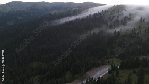 Clouds move over the forest and road in mountains. A view of mountains shows clouds moving over tall trees and a winding road in the early morning. This location has a mixture of greenery and mist rol