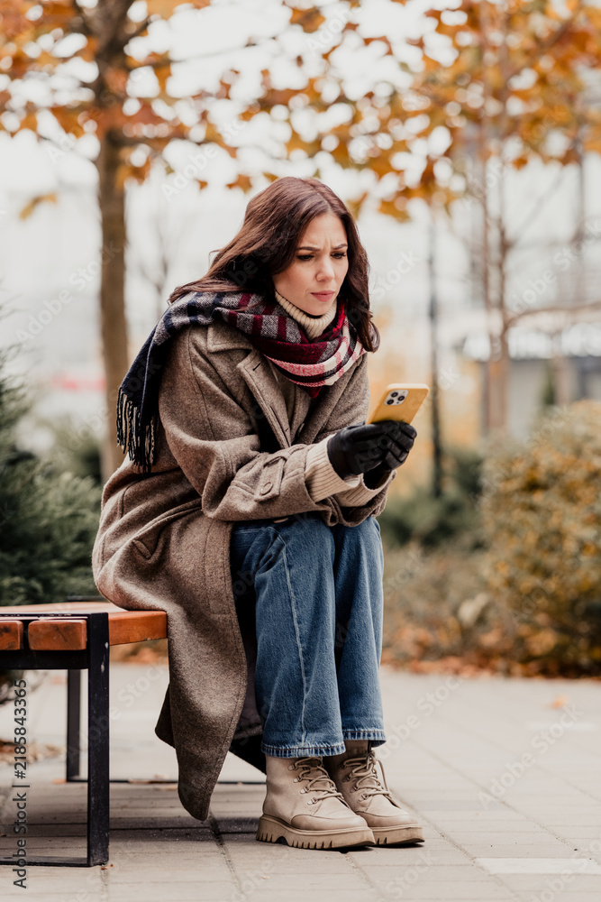 Obraz premium Young woman in coat and scarf sitting on a bench in autumn checking her phone with a worried expression