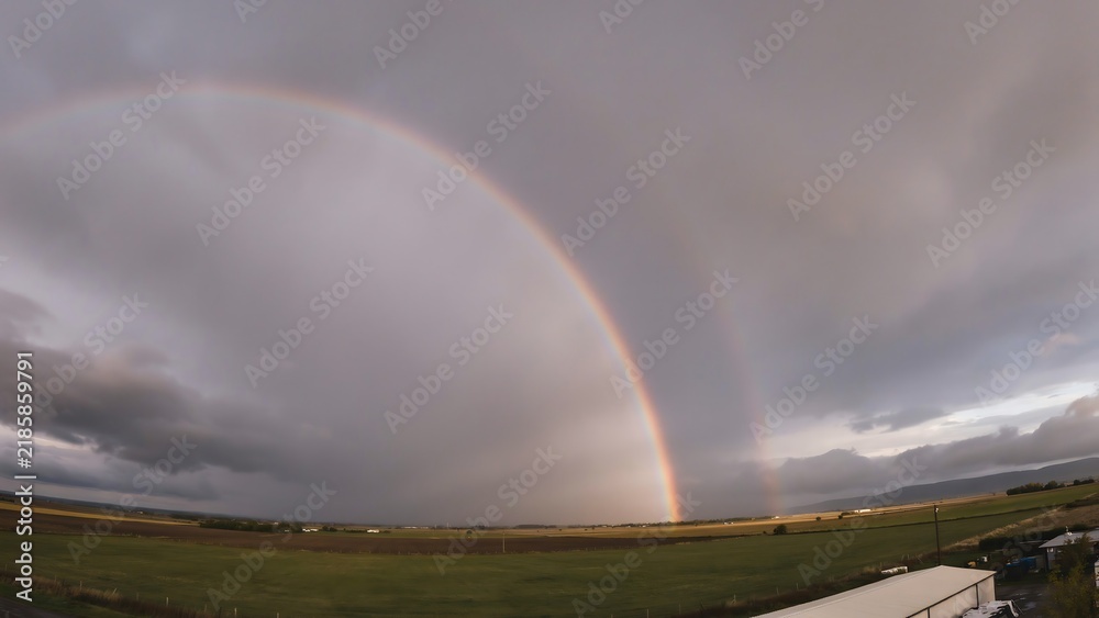 Obraz premium Rainbow arcs across a stormy sky over a wide green field.