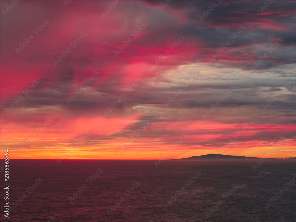 Fototapeta premium Intense crimson and orange sunset clouds spread across the sky above the Pacific Ocean off the Southern California coast, creating a dramatic fiery atmosphere over the distant shoreline and horizon.