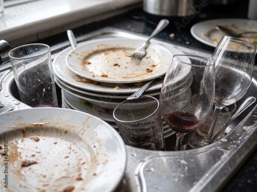 A cluttered stainless steel kitchen sink filled with stacks of unwashed plates, dirty glasses, and used cutlery waiting to be cleaned after a meal