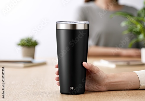Hand holding a sleek, black insulated tumbler with a silver rim on a desk