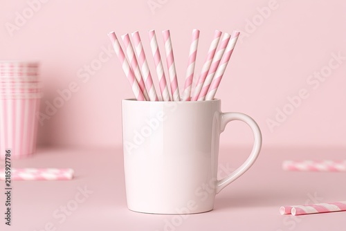 A white mug filled with pink and white striped straws, on a pink background