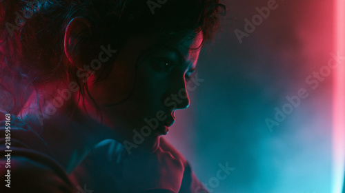 Close-up portrait of a thoughtful young woman in futuristic cyberpunk setting with dramatic neon pink and blue lighting, moody atmosphere, and hazy smoke. Cinematic sci-fi concept.