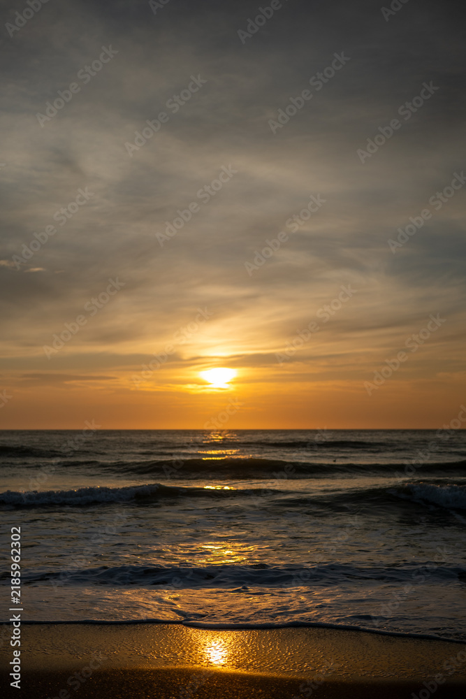 Obraz premium golden sunrise over sea horizon with wave reflections, vertical shot of ocean sunset with sun path on wet sand, scenic coastal sunrise with overcast sky and glowing water