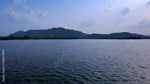 Udaipur, Rajasthan, India: A green mountain ridge stretches across the horizon beyond Lake Pichola's rippling water under a pale blue sky.