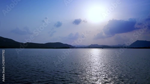 Udaipur, Rajasthan, India: Bright sunlight breaks through clouds and reflects across Lake Pichola, with dark hill silhouettes along the horizon.