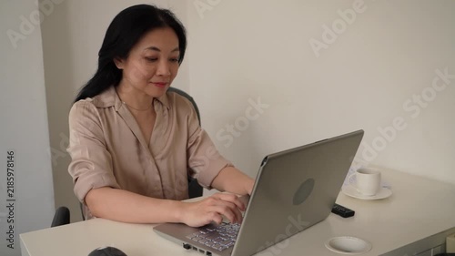 Positive Asian professional woman smiles gently while typing on a laptop at an office desk, representing productivity, confidence and job satisfaction.