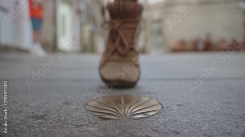 Close-up, low-angle perspective of a bronze scallop shell marker set into a stone pathway, with the out-of-focus legs and feet of a pedestrian walking in the background.