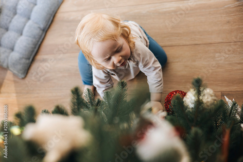 Happy little toddler decorating a Christmas tree at home, viewed from above. Cute child reaching for festive ornaments during Christmas preparation, enjoying a cozy family holiday tradition.