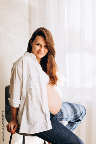 Pregnant woman sitting on a chair by a bright window, smiling at the camera and showing her baby bump. Warm maternity portrait representing pregnancy, motherhood and anticipation.