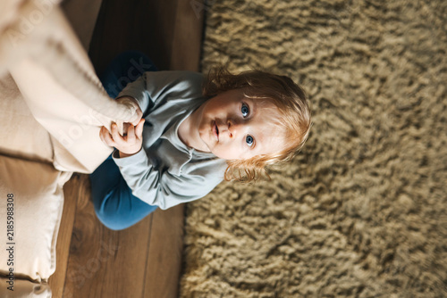 Cute little toddler sitting on the floor at home, holding soft fabric and looking up at the camera.Natural lifestyle portrait of a curious child exploring the home environment, representing childhood