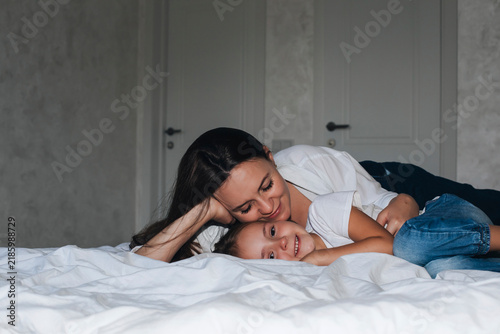 Loving mother and young daughter lying together on a bed, smiling and cuddling at home. Warm family moment expressing maternal love, affection, bonding, comfort and happy childhood.