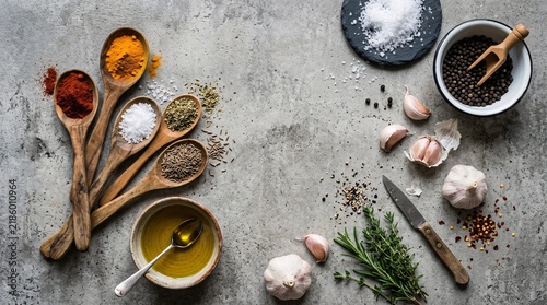 Culinary flat lay on grey concrete with wooden spoons of colorful spices, sea salt, garlic cloves and a small bowl of golden olive oil