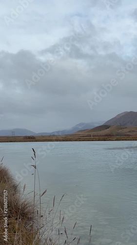 Calm river or canal landscape with reed-edged shore, expansive horizon, distant hills and mountains under cloudy sky, tranquil wide nature scene for travel and outdoors