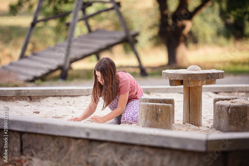 фотографія Young girl sitting in an outdoor sandbox digging into the sand during summer pla