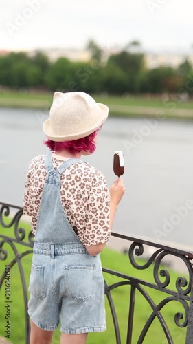 Rear view of child with short pink hair wearing straw hat, floral shirt, and denim overalls while holding partially bitten chocolate coated ice cream bar at riverside railing, soft overcast light