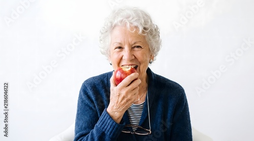 An elderly woman is eating a fresh red apple; isolated against a white studio background. AI generated.