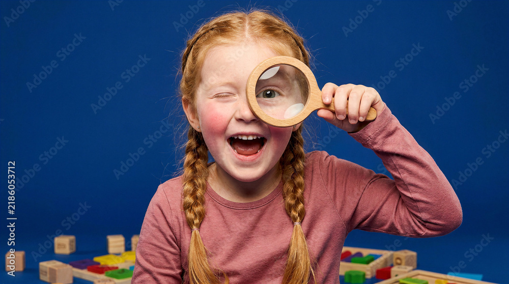 Fototapeta premium Curious young girl with red braids peers through a magnifying glass