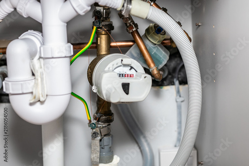 Foto Under-sink domestic water meter assembly with copper and brass supply lines, a white plastic meter or control module housing, and waste trap assembly