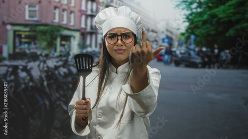 Фотография Chef woman in white chef hat and uniform holds spatula and shows middle finger on street with parked bikes and storefronts; defiance boldness