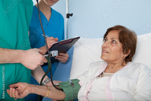 Doctor or male nurse checking blood pressure of senior female patient in hospital bed while nurse takes notes on clipboard.