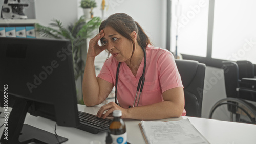 Quadro su tela Woman doctor in pink scrubs types on computer with stethoscope, sorts papers and puts hand to forehead for headache in building; fatigue